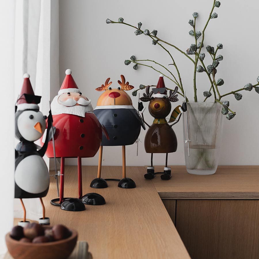 Christiania 1739 Julekolleksjon Figur Reinsdyr 18cm, reinsdyrfigur står sammen med flere juledekorasjoner, inkludert en pingvin og en julenisse, på et stilrent bord.