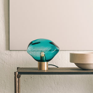 Hadeland Glassverk Crystal Stone bordlampe i fargen Blågrønn på en hylleseksjon, som gir stemningsfull belysning og elegant interiørdesign.