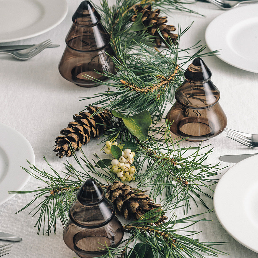 Hadeland Glassverk Gran Juletre Mørk Kakao utstilt i en dekorsetting med granbar og kongler, tilføyer naturlig eleganse til borddekkingen.
