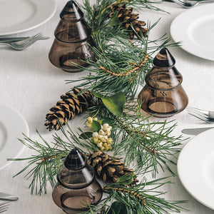 Hadeland Glassverk Gran Juletre Mørk Kakao utstilt i en dekorsetting med granbar og kongler, tilføyer naturlig eleganse til borddekkingen.