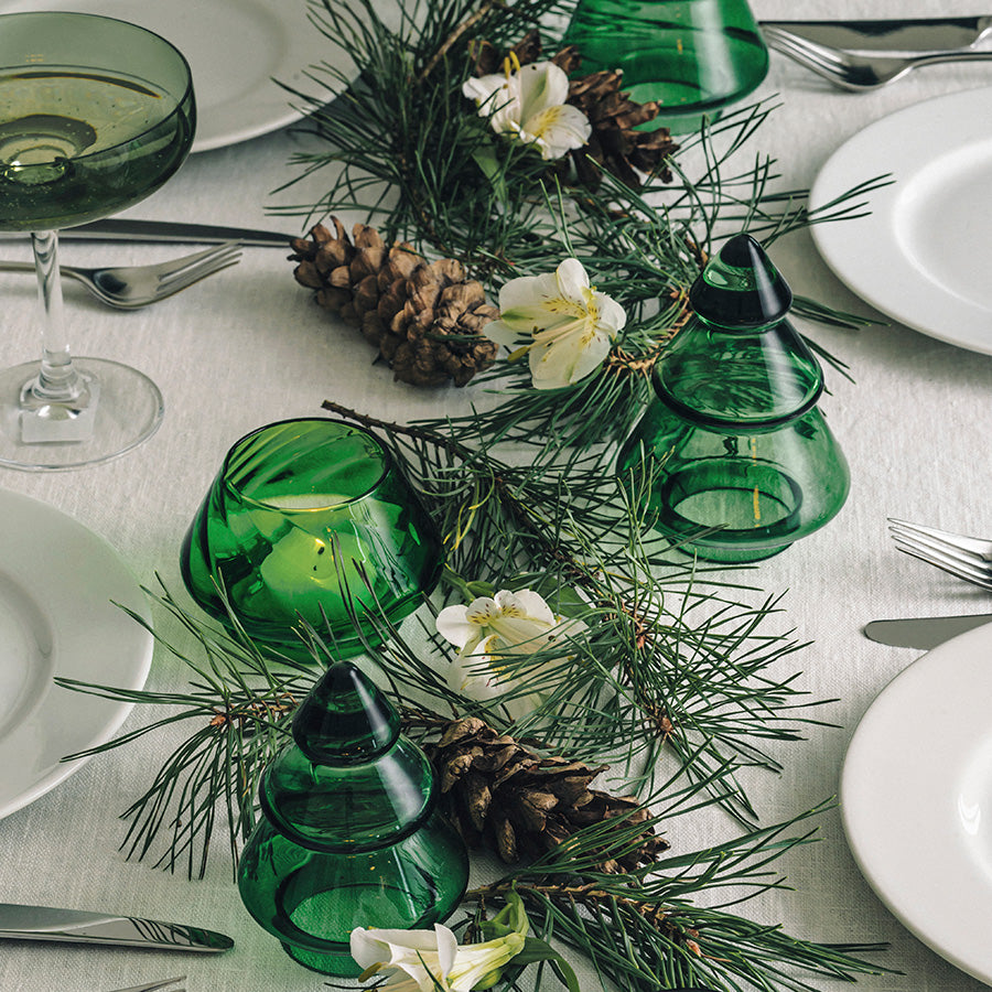 Dekorativ borddekorasjon med Hadeland Glassverk Gran Juletre 9,8 cm i vintergrønn glass omgitt av naturlige elementer.