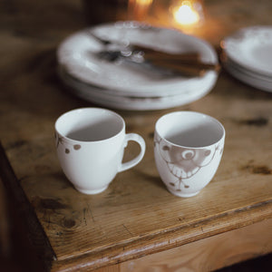 To Porsgrund Porselænsfabrik Maxistrå Krus 35cl Nomi Beige plassert på et trebord sammen med tallerkener, passer perfekt til hyggelige stunder.