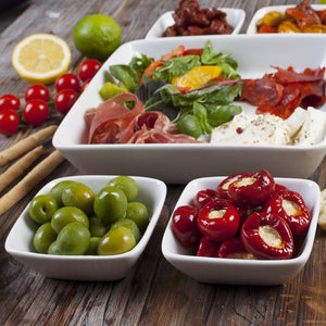 Porsgrunds Porselænsfabrik Sense Firkant Bolle 10 cl, viste sammen med en rekke ferske ingredienser som grønne og røde oliven, skinke og salat på en rustikk treoverflate.