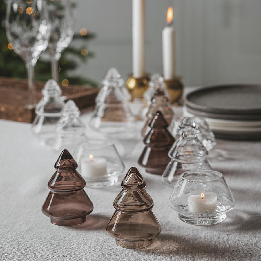 Nærbilde av Hadeland Glassverk Gran Juletre Mørk Kakao 11,5cm sammen med andre glassfigurer, skaper en koselig atmosfære med lys og skygger.