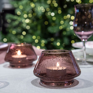 Hadeland Glassverk to rosa lyslykter med tent lys, vakkert presentert på bord med grønn bakgrunn og stemningsfull belysning.