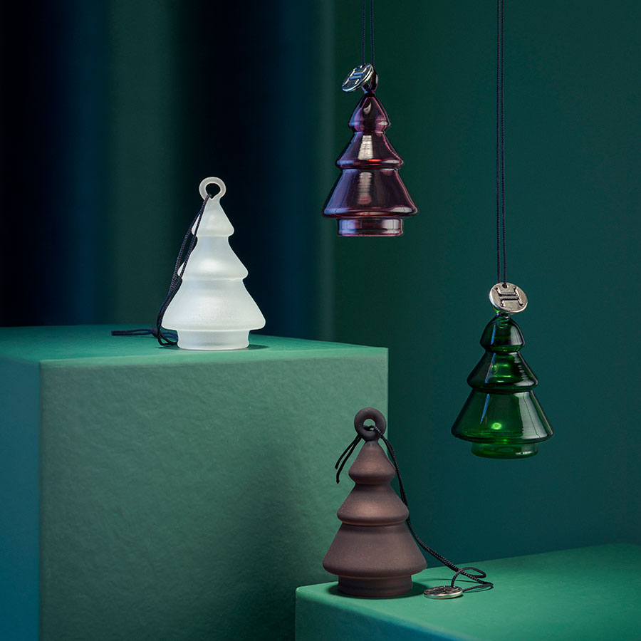 Hadeland Glassverk juletrepynt i forskjellige farger, inkludert grønne, hvite og burgunder grantrær som står på fargede plater.