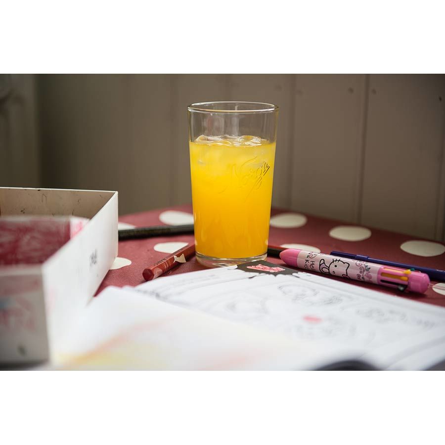 Norgesglasset drikkeglass 400ml med en gul drink, plassert ved siden av fargeleggingsutstyr på et bord.