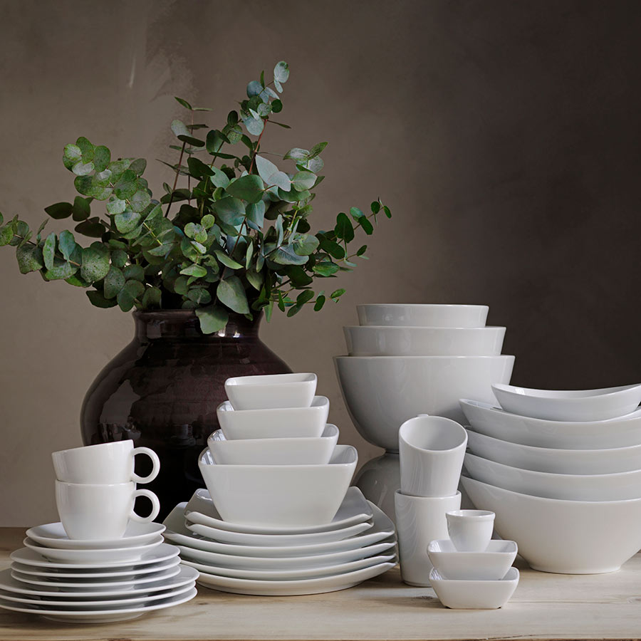 Utvalg av hvite keramiske tallerkener og skåler fra Porsgrunds Porselænsfabrik, inkludert Porsgrunds Porselænsfabrik Sense Krus Maud m/Hank, presentert med grønne planter.