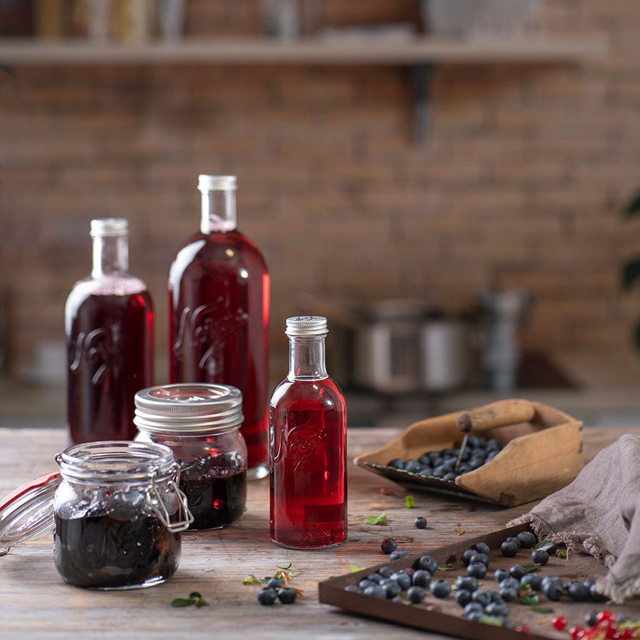 Norgesglass flasker fylt med rød drikk laget av bær på et bord, sammen med glasskrukker, som viser bruken av Norgesglasset til saft og stille drikker.