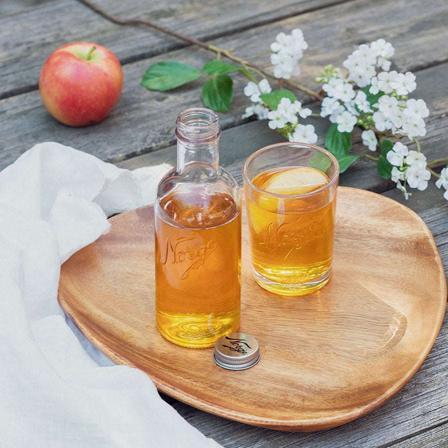 Norgesglass flaske med gyllen drikk servert i et glass, plassert på en treplate sammen med eple og blomster, illustrerer bruken av Norgesglasset i servering.