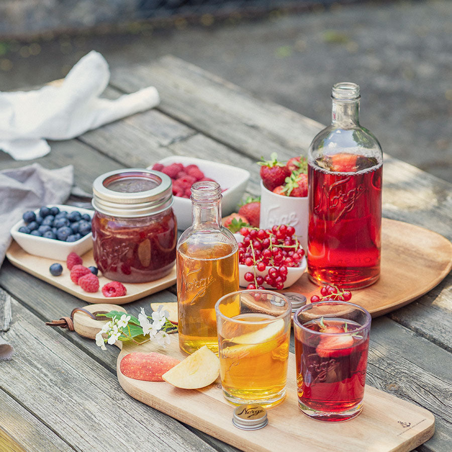 Samling av Norgesglasset flasker med fargerike drikker og frukt, vist på et bord med treplater, fremhever Norgesglassets stil og allsidighet for servering.