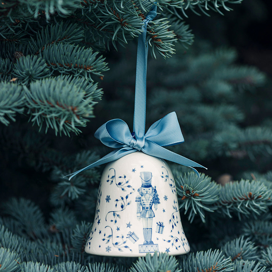 Porsgrunds Porselænsfabrik PP Jul Ballerina Klokke Hengepynt 8cm Blå, dekorativ klokke med blått bånd hengende i et juletre med grønne grener, ideell for juledekorasjoner.