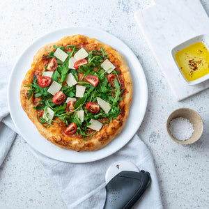 Pizza toppet med ruccola, cherrytomater og parmesanost servert på Porsgrunds Porselænsfabrik Sense pizzafat, ved siden av en liten skål med olivenolje.