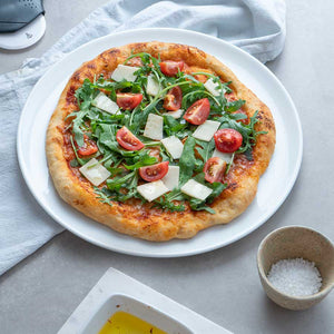 En pizza dekorert med ruccola og cherrytomater på Porsgrunds Porselænsfabrik Sense pizzafat, omringet av en lysegrå overflate.