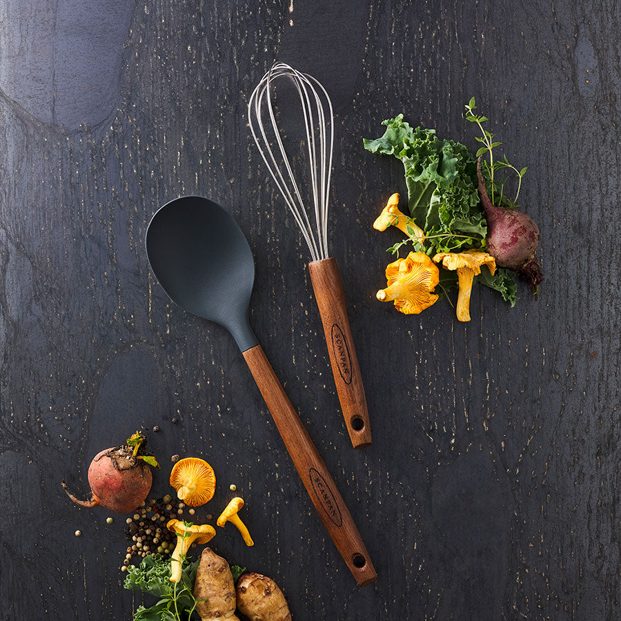 Scanpan Serveringsskje 31cm Karbonisert Ask sammen med en visp og grønnsaker, illustrerer det elegante og funksjonelle kjøkkenutstyret.