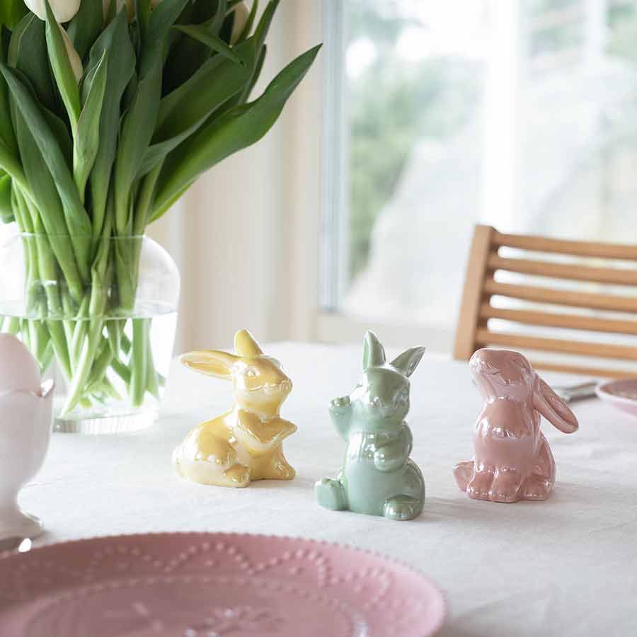 Tre keramiske harer i rosa, grønn og gul farge plassert på et bord dekket med duk. Blomster i en vase står i bakgrunnen, noe som gir en våraktig stemning.
