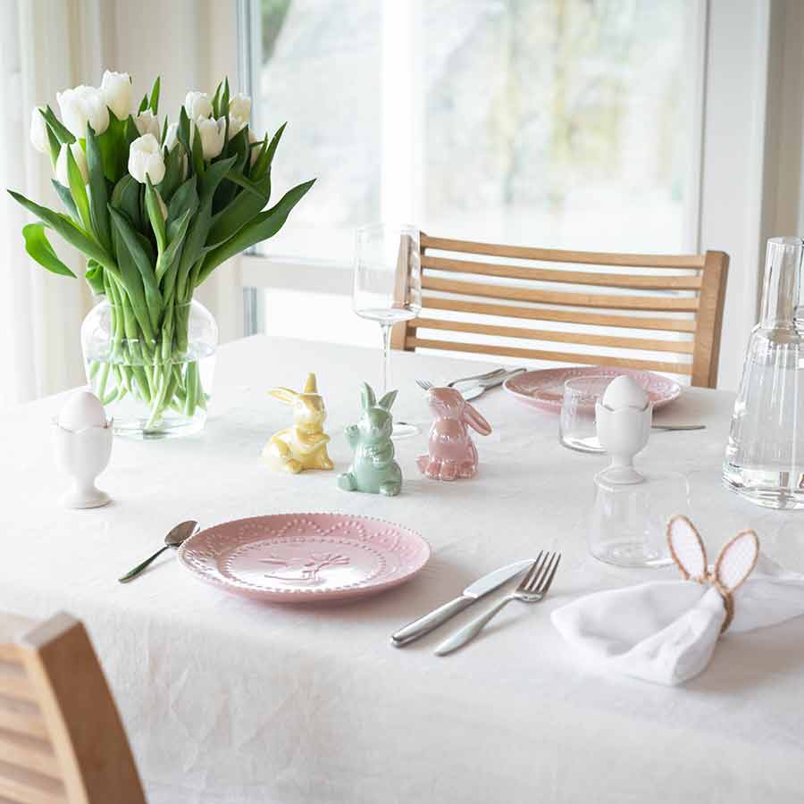 Et bord med tre keramiske harer i pastellfarger - rosa, grønn og gul - sammen med en hvit eggeholder og en tulipanbukett i en glassvase, skaper en innbydende påskedekor.