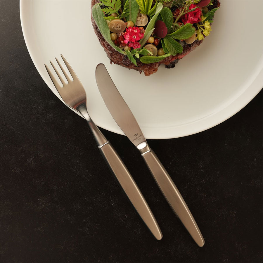 Skaugum bestikksett med gaffel og kniv på en mørk tallerken med gourmetmat. Designet er tidløst og passer enhver middag.