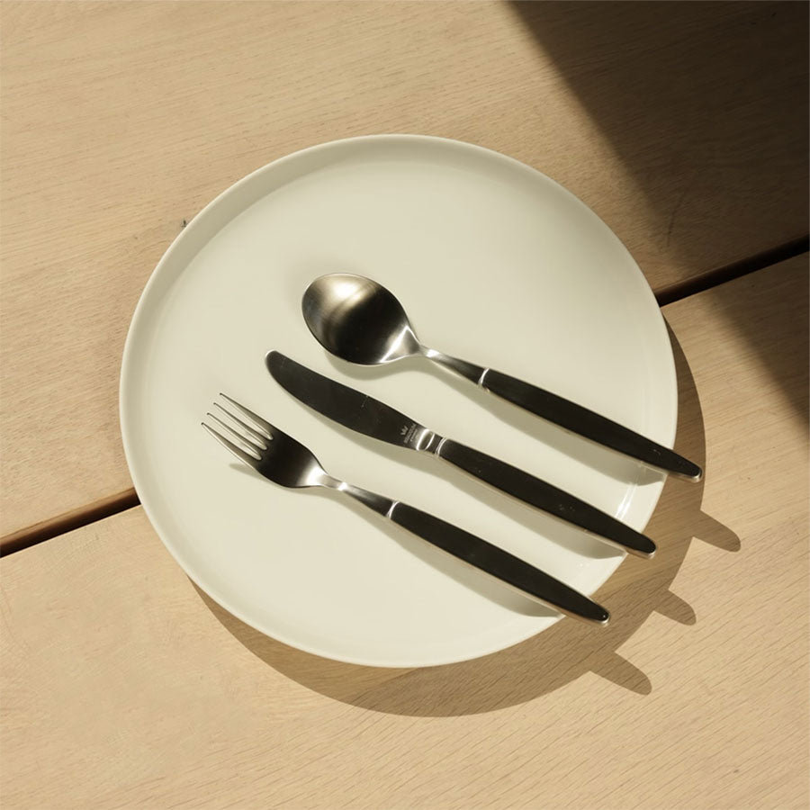 Børstet stål Skaugum bestikksett presentert på en hvit tallerken med spiseskje, gaffel og kniv. Ideelt for stilige borddekorasjoner.