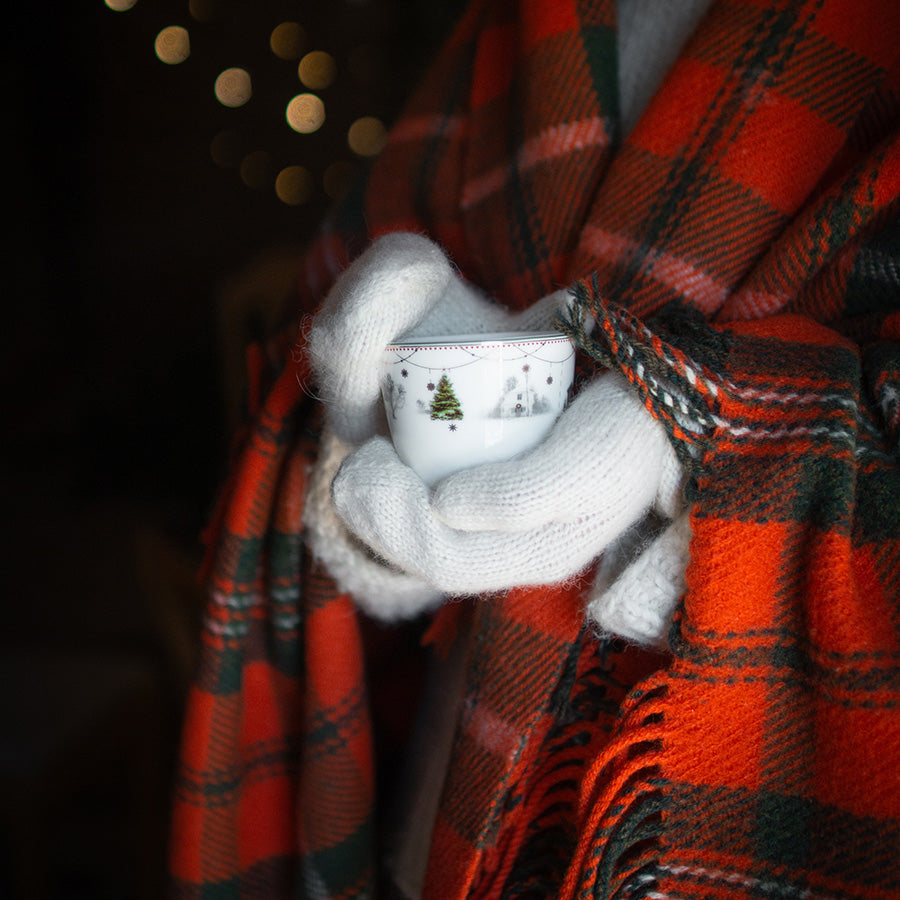 En person holder en julekopp fra Wik & Walsøe i et teppe, med lunt lys i bakgrunnen, som symboliserer julens hygge.