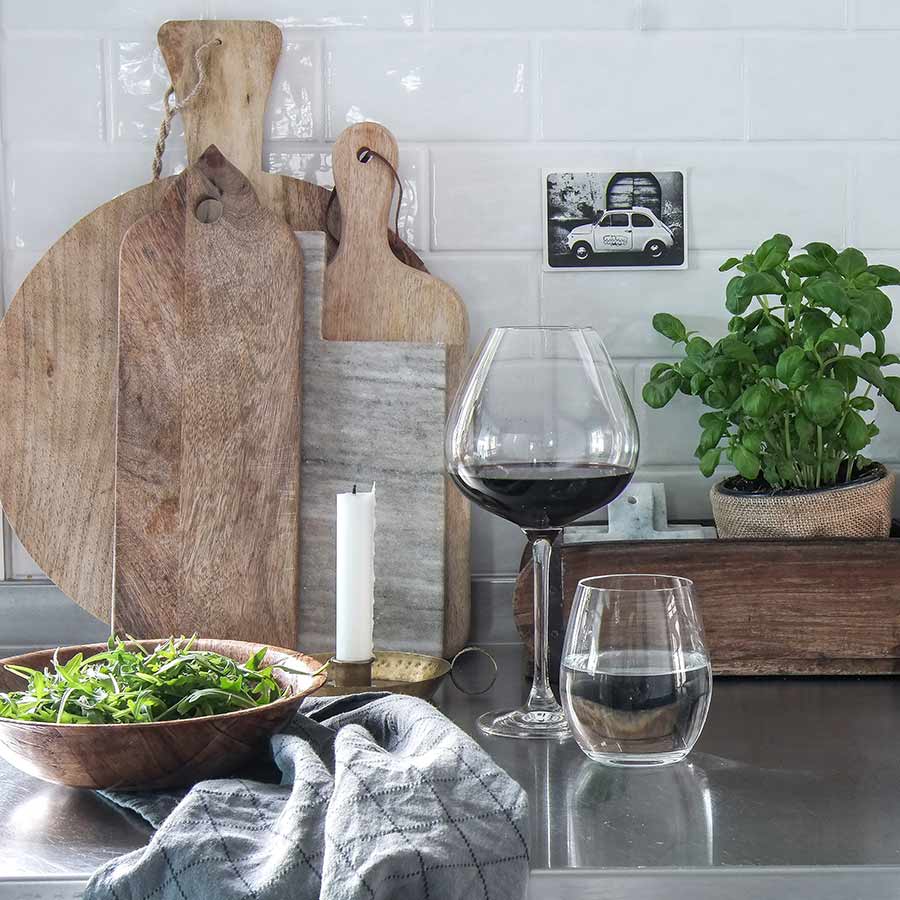 Stilig kjøkkenoppsett med Hadeland Glassverk rødvinsglass, en stor salatbolle og et lite glass med vann. Beige og brune skjærebrett i bakgrunnen, samt grønn basilikumplante. Perfekt for å skape en innbydende atmosfære.