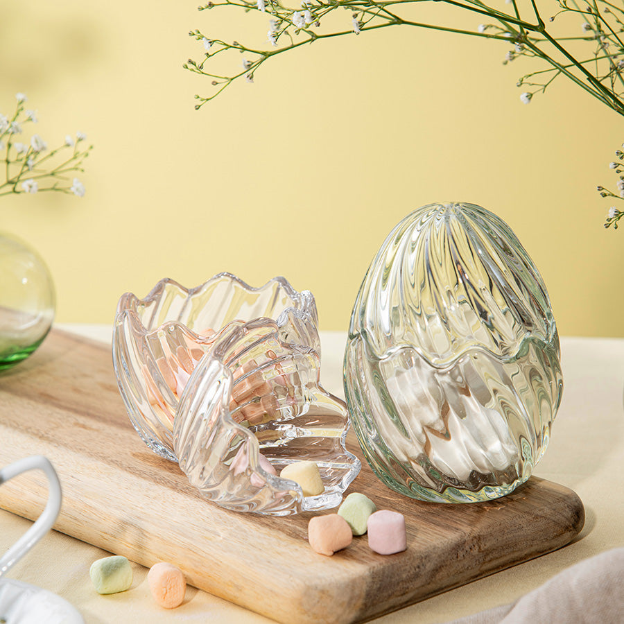 En elegant glasspåskeegg med en liten hare på toppen, plassert på et dekket bord med drikkeglass og grønne planter. Eggformet design i frostet glass som gir en dekorativ detalj til borddekorasjonen.