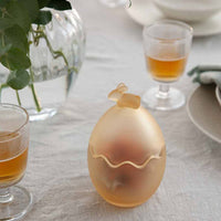 En elegant glasspåskeegg med en liten hare på toppen, plassert på et dekket bord med drikkeglass og grønne planter. Eggformet design i frostet glass som gir en dekorativ detalj til borddekorasjonen.