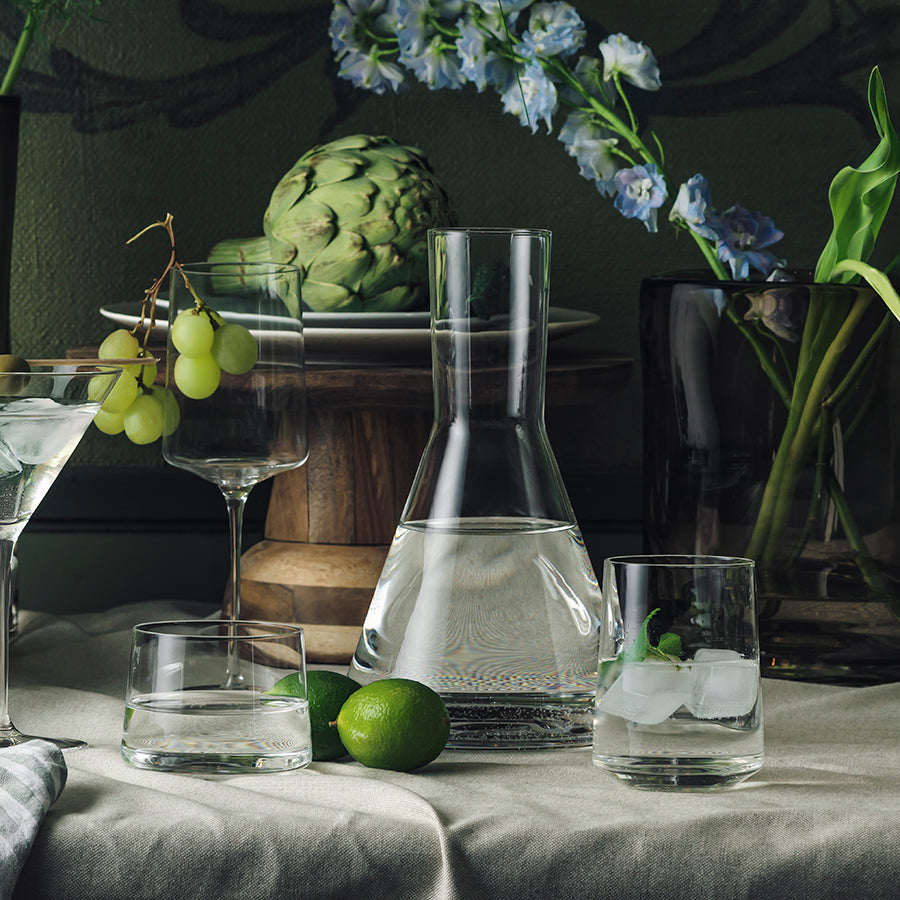 Samling av Amalfi glass fra Hadeland Glassverk, inkludert karaffel, longdrinkglass, og vannglass, dekorert med friske grønnsaker og blomster for en elegant setting.