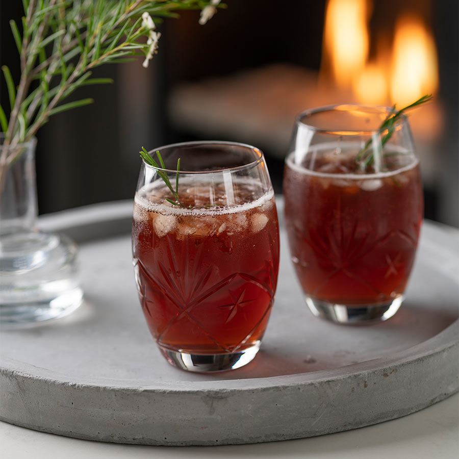 To munnblåste Hadeland Glassverk Finn vannglass på et grått fat, fylt med brus og pyntet med rosmarin. I bakgrunnen skimtes en koselig peis med flammer.