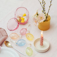 Et vakkert dekket bord med påskeegg i glass, inkludert en glasskål med lokk fylt med fargerike godterier, en vase med blomster og et tent lys. Bordet har også en gullskje og servietter, som skaper en festlig stemning for påske.