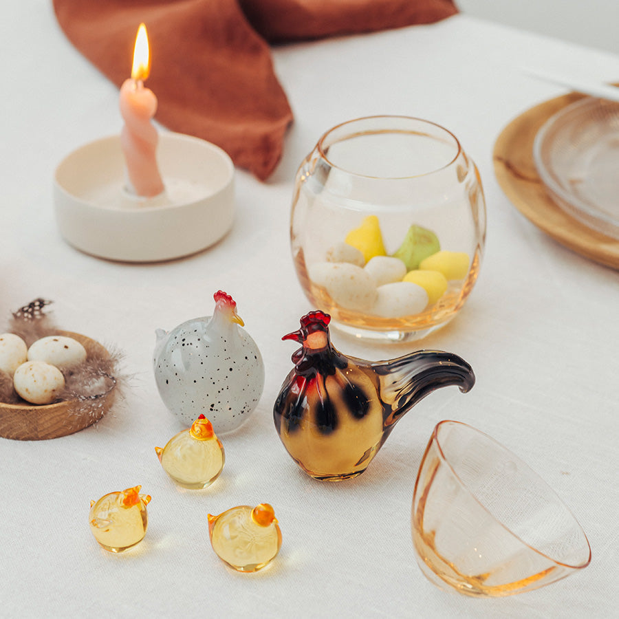 Tre dekorative glasshøns fra Hadeland Glassverk i gult og en hvit glassfigur med prikker. I bakgrunnen er det blomster i en gul vase og en delikat duk på bordet, som bidrar til en påskestemning.