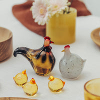 Tre dekorative glasshøns fra Hadeland Glassverk i gult og en hvit glassfigur med prikker. I bakgrunnen er det blomster i en gul vase og en delikat duk på bordet, som bidrar til en påskestemning.
