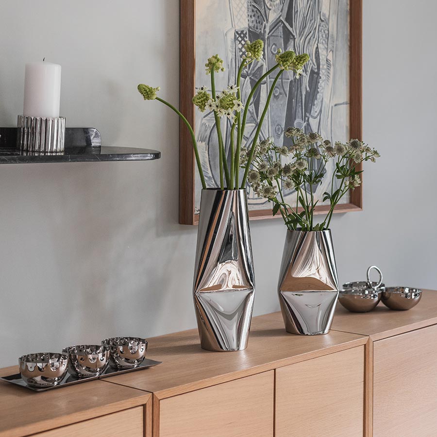 Løve vaser i rustfritt stål med moderne design, plassert på en trekommode med blomsterdekorasjoner. Perfekte som stilige vaser til stue eller spisebord.