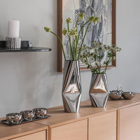 Løve vaser i rustfritt stål med moderne design, plassert på en trekommode med blomsterdekorasjoner. Perfekte som stilige vaser til stue eller spisebord.