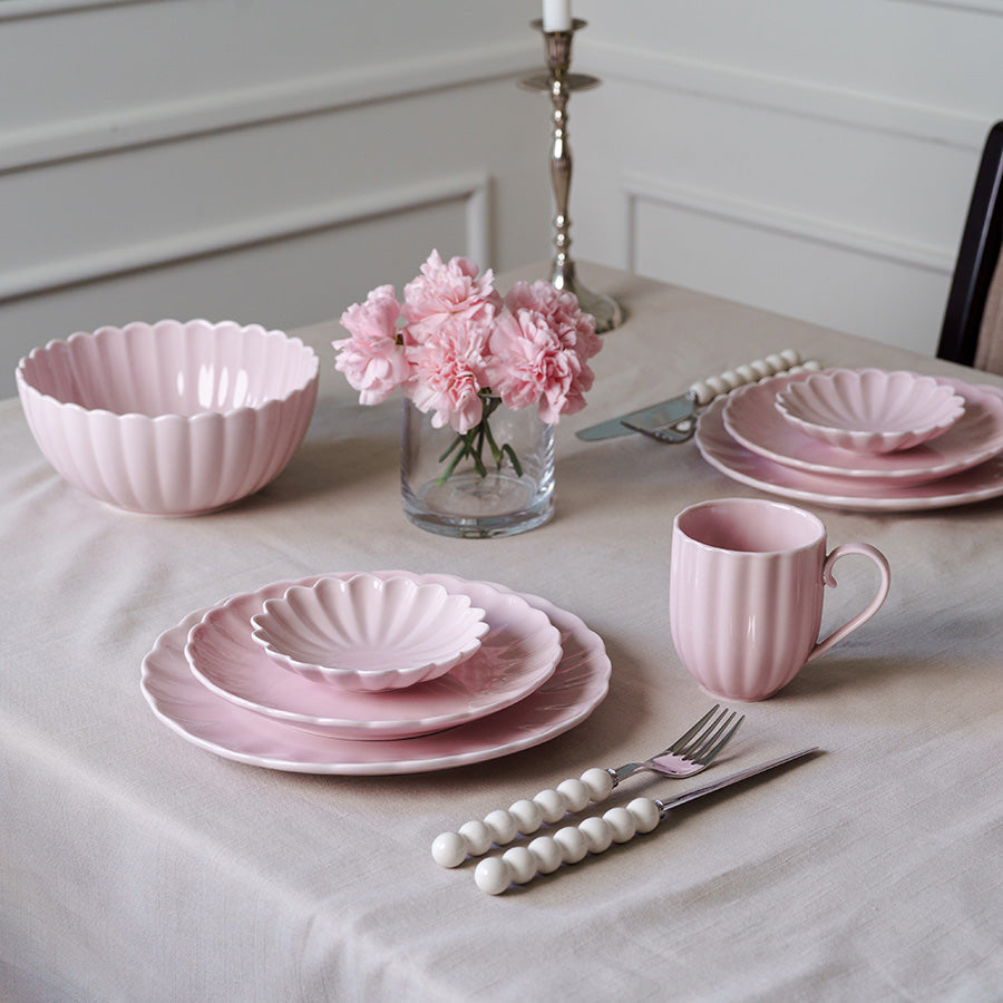En elegant borddekking med rosa tallerkener, skåler og krus i en delikat atmosfære. Bordet er dekket med en lys duk, blomster i vase og stilige bestikk med perle-design.