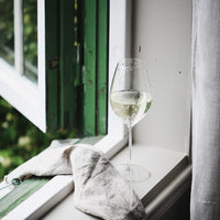 Riedel vinglass med hvitvin plassert på vinduskarmen, omgitt av grønn vindusramme og et belegg av naturlig lys.