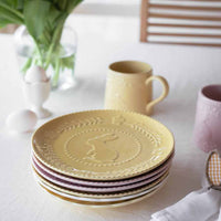Fargerike tallerkener og krus i påskedesign, inkludert en gul tallerken med kaninmotiv øverst, stablet på et bord med påskeegg, blomster og servietter.