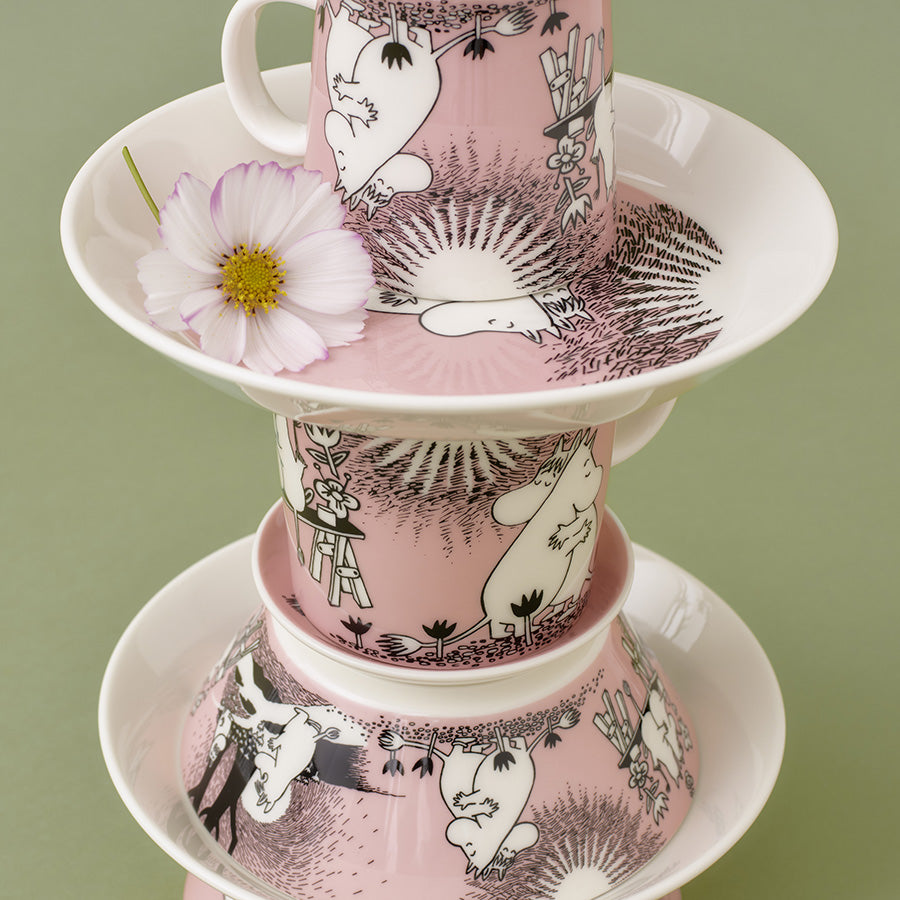 Moomin Arabia asjett stablet med kopper på toppen, dekorert med illustrasjoner av Mummitrollet, på en grønn bakgrunn. Perfekt for borddekking.