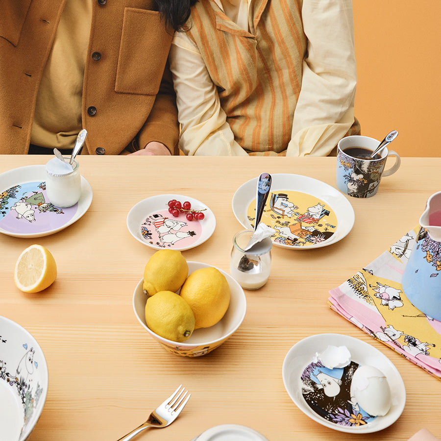 To personer som deler et måltid med Moomin Arabia Mummi asjett 15cm Familietid, omgitt av smakfulle retter og levende farger.