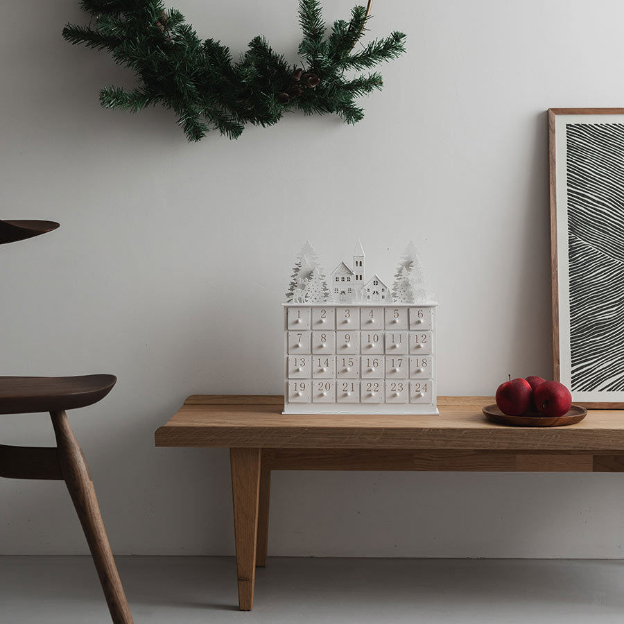 Christiania 1739 Julekolleksjon Julekalender I Tre på en trebord, dekorert med juledekorasjoner og røde epler, perfekt for adventstiden.