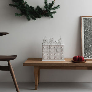 Christiania 1739 Julekolleksjon Julekalender I Tre på en trebord, dekorert med juledekorasjoner og røde epler, perfekt for adventstiden.