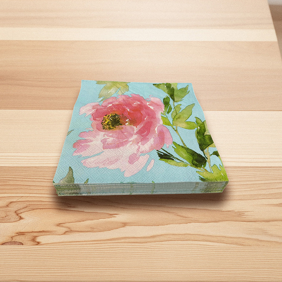Servietter 33x33 cm med rosa pionmotiv, blå bakgrunn og grønne blader, stablet på et lyst trebord