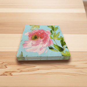 Servietter 33x33 cm med rosa pionmotiv, blå bakgrunn og grønne blader, stablet på et lyst trebord
