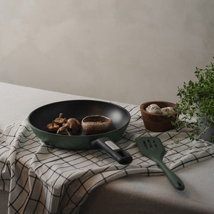 Christiania 1739 Plus Stekepanne 28cm Grønn med sopp i pannen og en spatula, plassert på et tekstil bord.