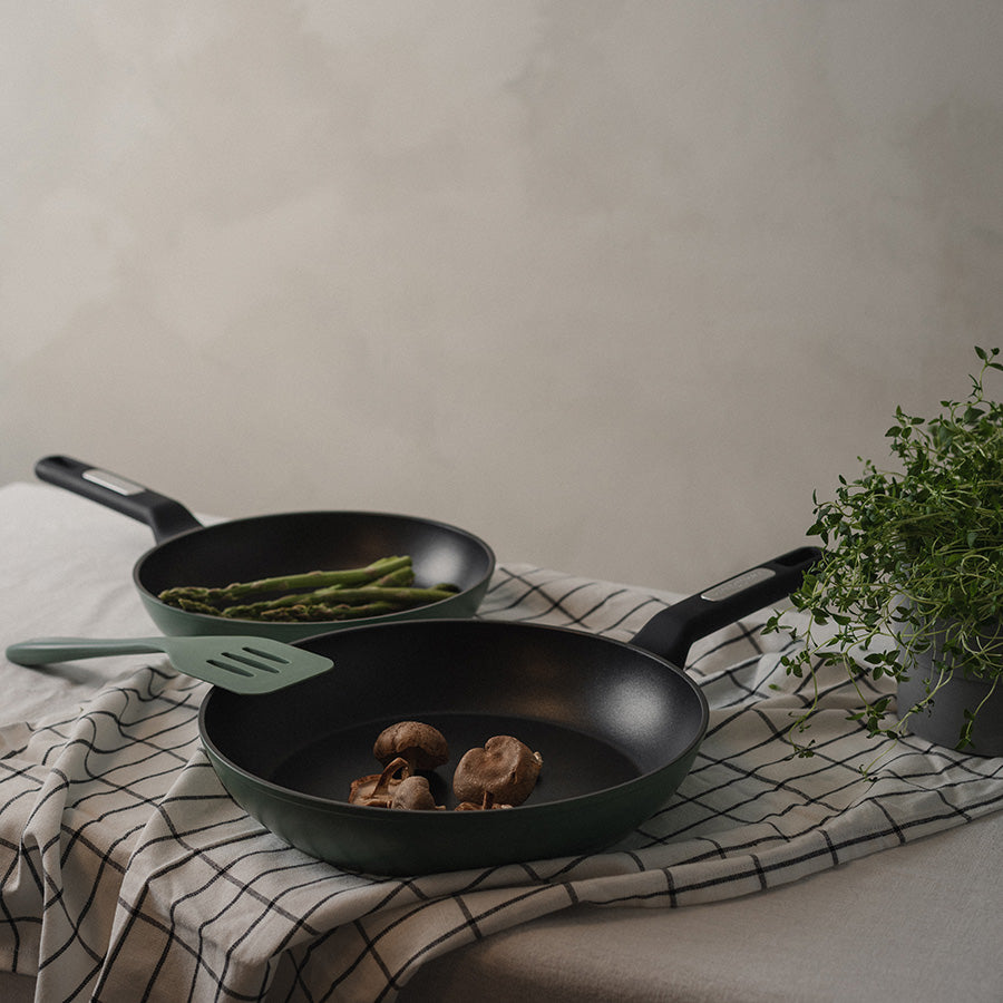 To Christiania 1739 Plus Stekepanner 28cm Grønn på et dekket bord, med grønnsaker som asparges og sopp.