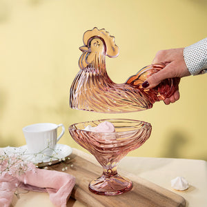 Hånd som løfter lokket av en rosa glasshane fra Hadeland Glassverk, som viser den funksjonelle og dekorative bruken til oppbevaring av søtsaker.