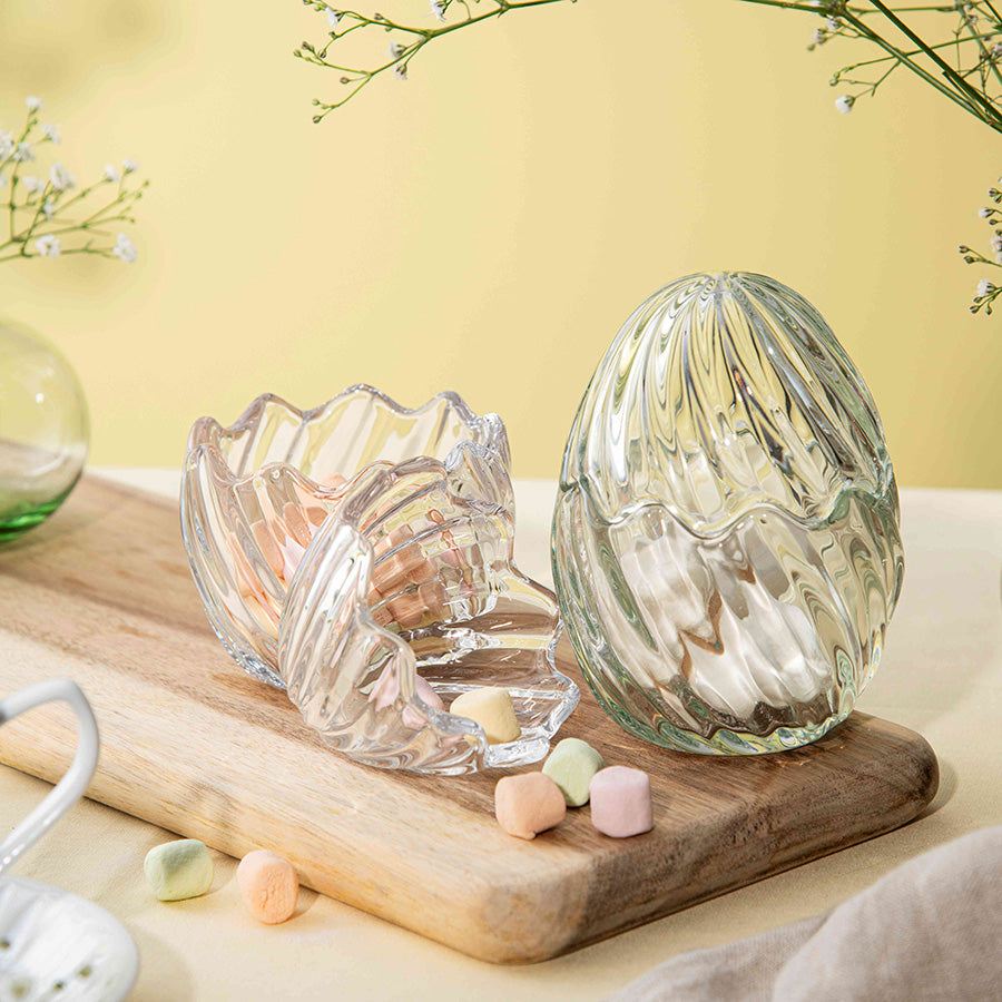 To dekorative glass påskeegg, ett grønt og et klart, fylt med fargerike godteri, plassert på et trebrett.