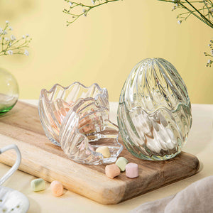 To dekorative glass påskeegg, ett grønt og et klart, fylt med fargerike godteri, plassert på et trebrett.