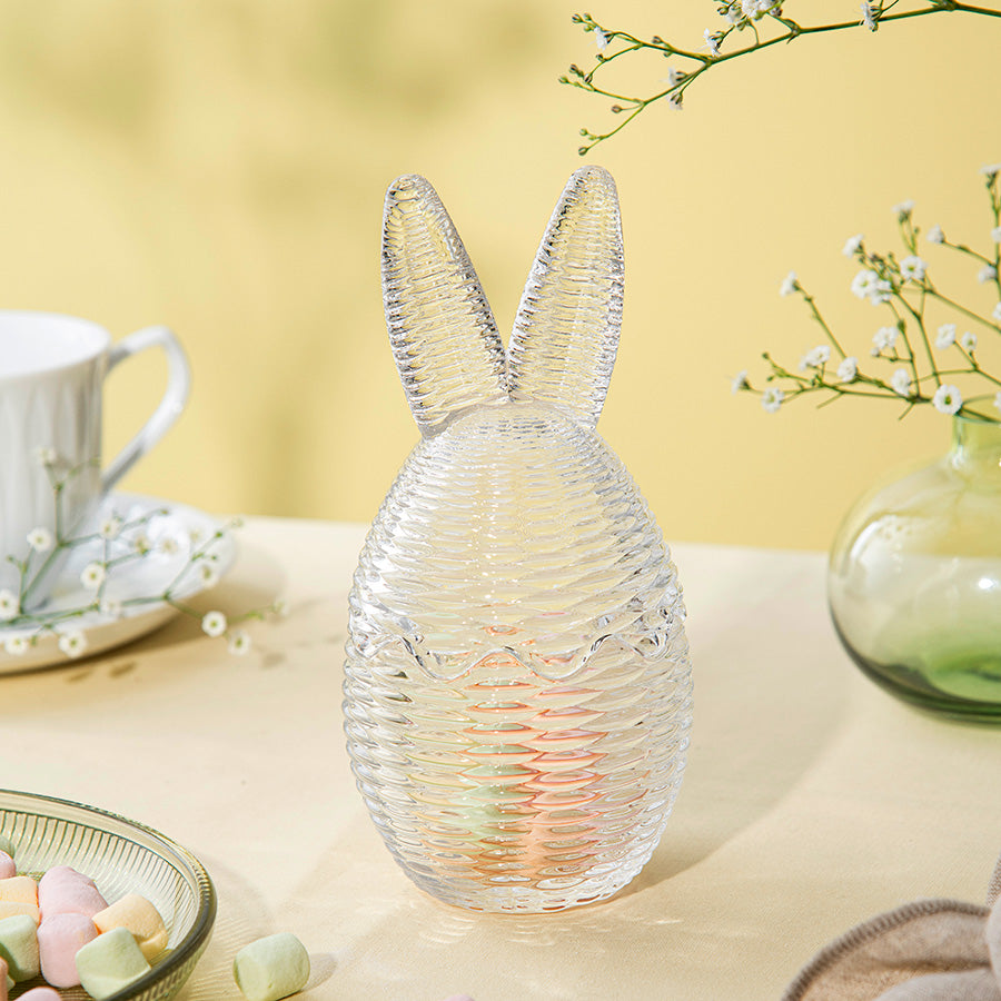 Hadeland Glassverk klart påskeegg med kaninører, designet i kurv-optikk, plassert på et bord med pastel godteri.