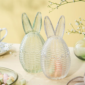 To Hadeland Glassverk påskeegg med kaninører i klart og grønt glass, dekorativt arrangert med søtsaker på et bord.